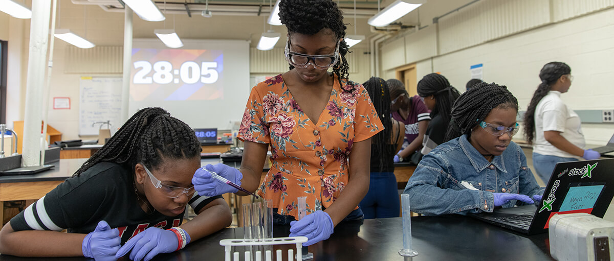 Three students looking at a test tub, adding droplets to the test tube and researching on a laptop